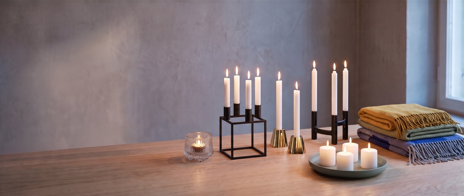 Lit candles in an Audo Kubus 4, Iittala Aalto brass candleholders, a Stelton Nordic Ora and a Blomus Vela, an Ultima Thule votive and folded Roros Tweed wool throws on an oak table