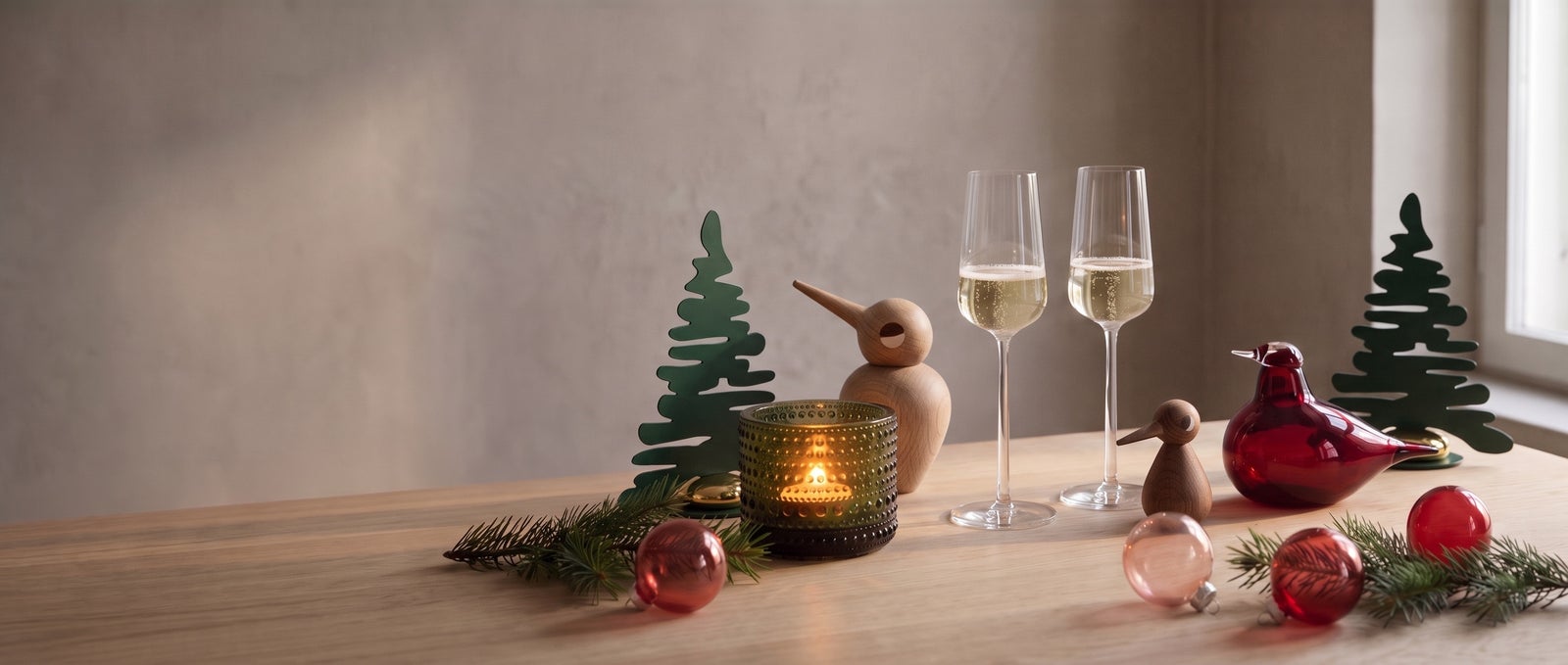 An Iittala Kastehelmi tealight, two Alessi Eugenia champagne flutes, a cranberry Toikka glass bird and red glass ornaments with two green Alessi Barkplace trees and two oak Architectmade birds on a long oak table