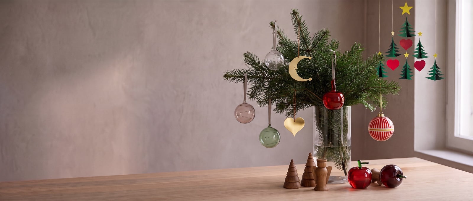 Cut fir branches in a glass vase hung with Iittala Sun Balls, Vitra Girard brass ornaments, Alessi Delight baubles and a glass apple, with Architectmade Jul trees on an oak table and a Flensted Christmas tree mobile by the window